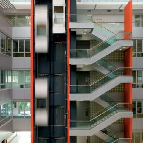 Widok na wewnętrzną klatkę schodową i szyby wind w nowoczesnym budynku, z przeszklonymi balustradami i jasnymi ścianami. Winda jest w ruchu, co widać po rozmytych drzwiach kabiny.